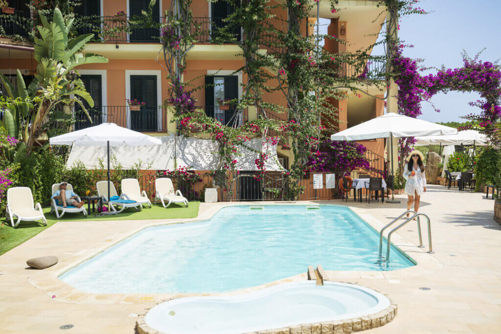 Galleria - Vista della piscina dell'Hotel Domus de Janas con una ragazza che cammina con un cocktail in mano ed un ragazzo che si riposa su un lettino sotto l'ombrellone