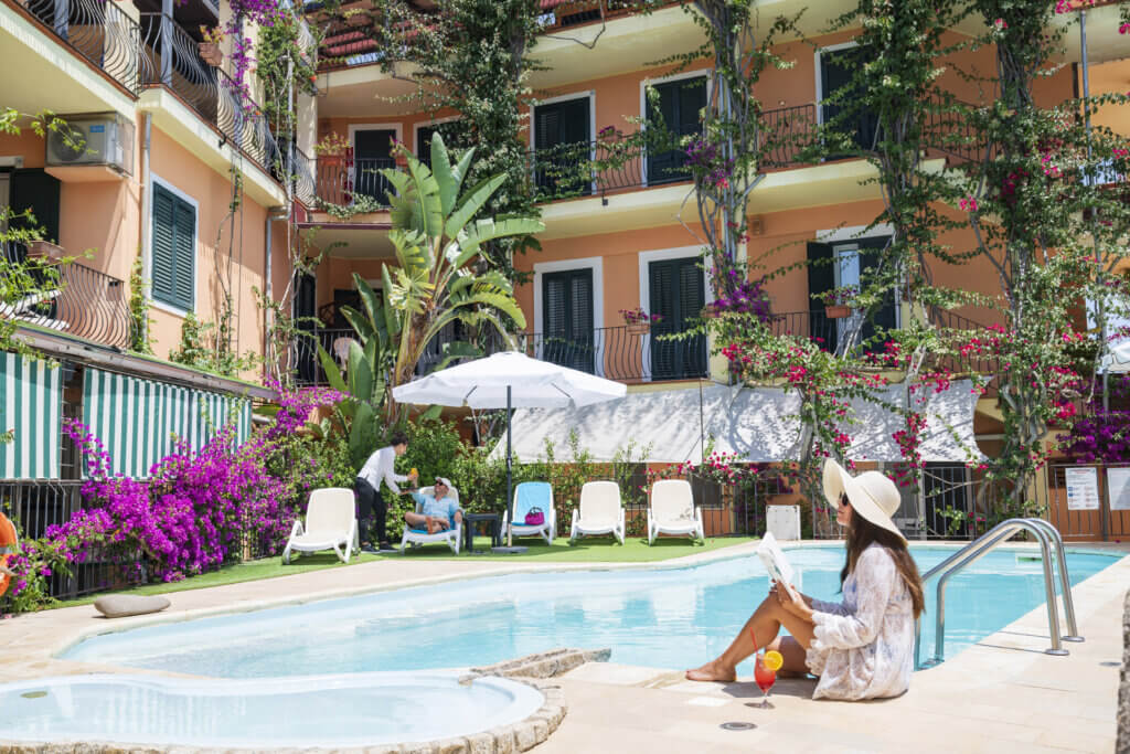 Galleria - La piscina dell'Hotel Domus de Janas con una ragazza che legge un libro con un cocktail vicino