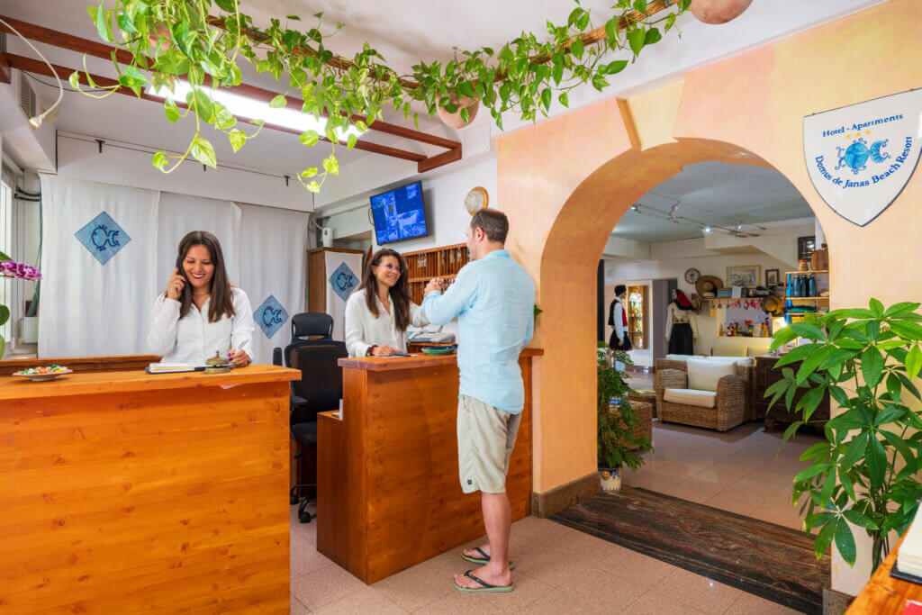 Galleria - La Reception dell'Hotel Domus de Janas conun cliente a cui vengono consegnate le chiavi della stanza mentre un'altra receptionist è al telefono