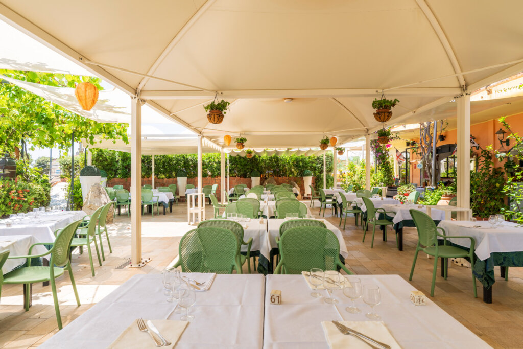 Galleria - La sala esterna del ristorante Il Pesciolino dell'Hotel Domus de Janas con tavoli apparecchiati ed ombrelloni