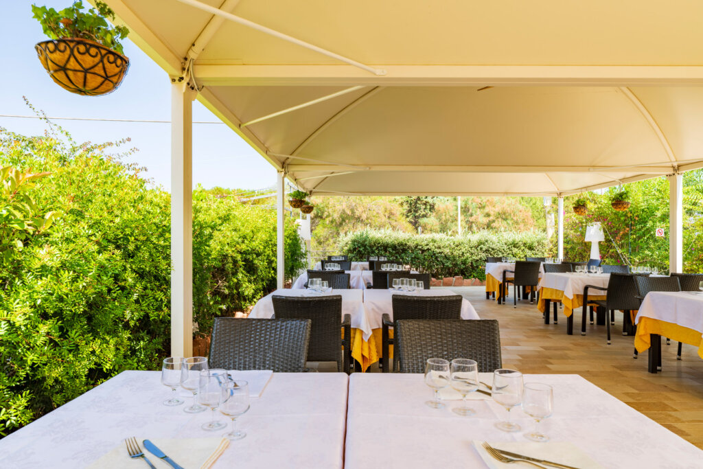 Galleria - La sala esterna del ristorante Il Pesciolino dell'Hotel Domus de Janas con tavoli apparecchiati ed ombrelloni