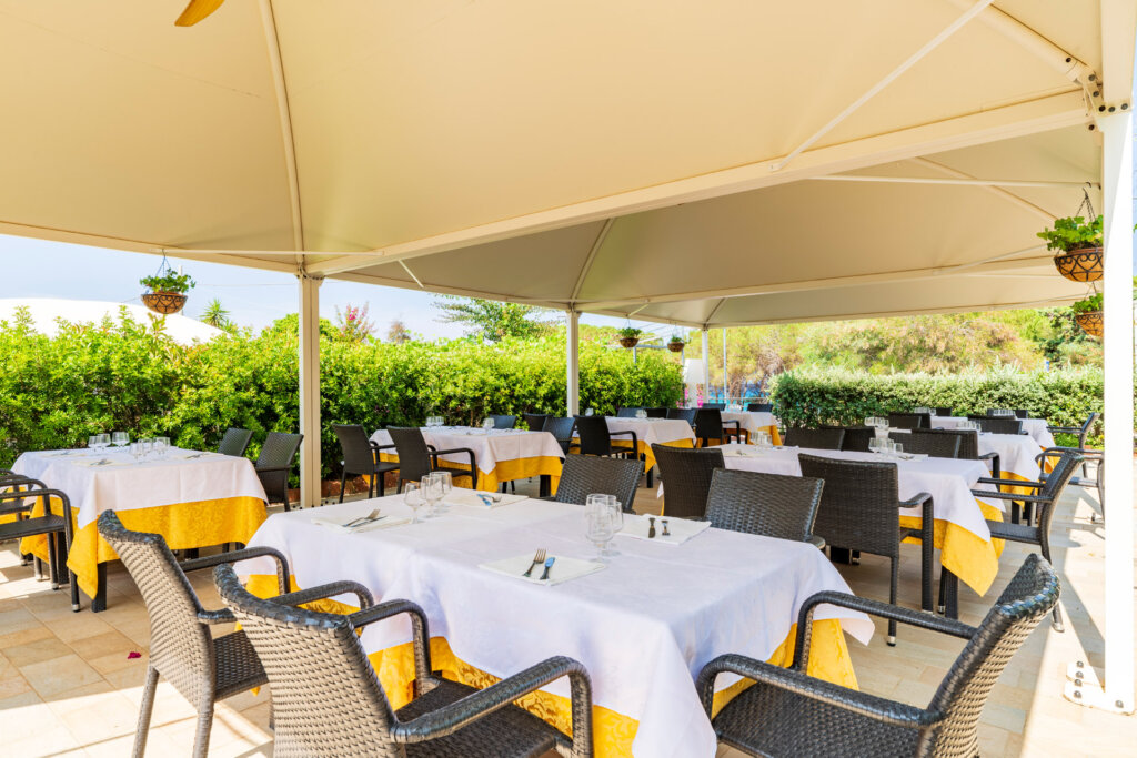 Galleria - La sala esterna del ristorante Il Pesciolino dell'Hotel Domus de Janas con tavoli apparecchiati ed ombrelloni